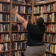 picture from the back of a black person with short black hair, a mask, and wearing dark clothes raising their hands in exclamation and joy when approaching the young adult section of a used bookstore